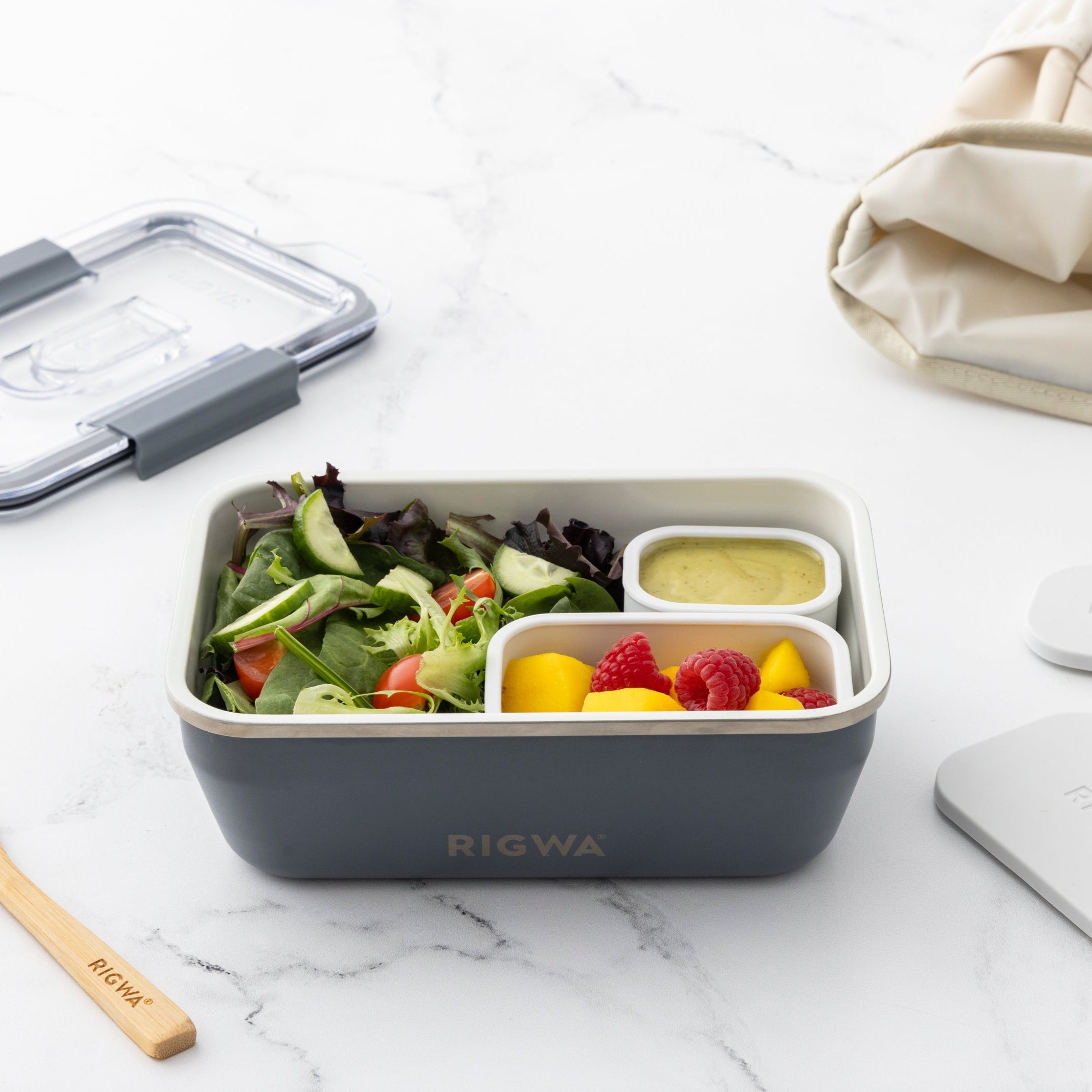 Bento box with salad and snacks on a marble surface, surrounded by a wooden fork and beige bag.
