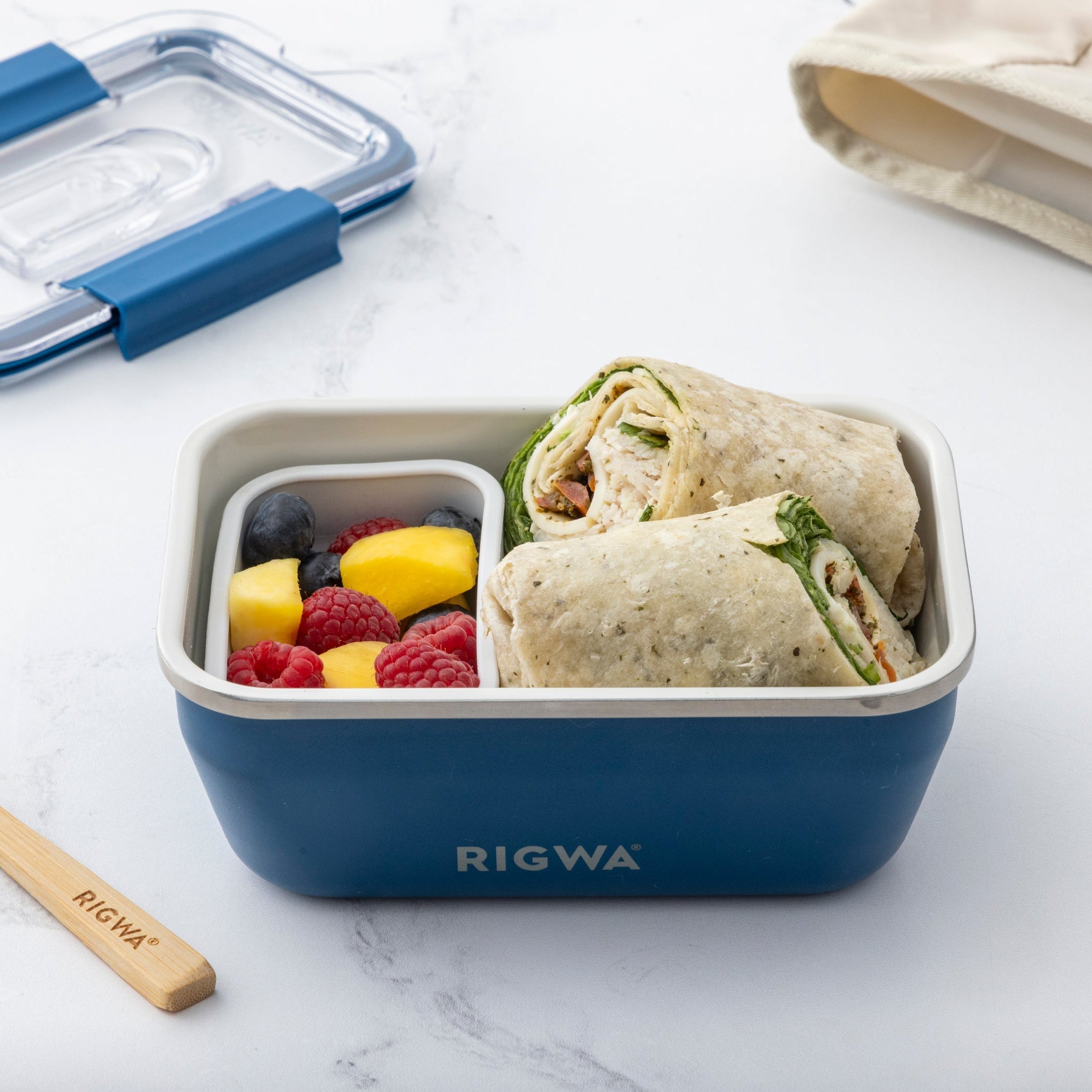 Blue lunch box with a wrap, fruits, and vegetables on a white surface with a beige bag and wooden fork.